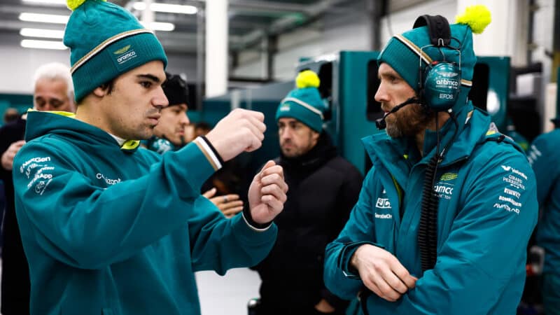 Lance Stroll raises his hands as if holding a steering wheel in the Aston Martin F1 pit garage