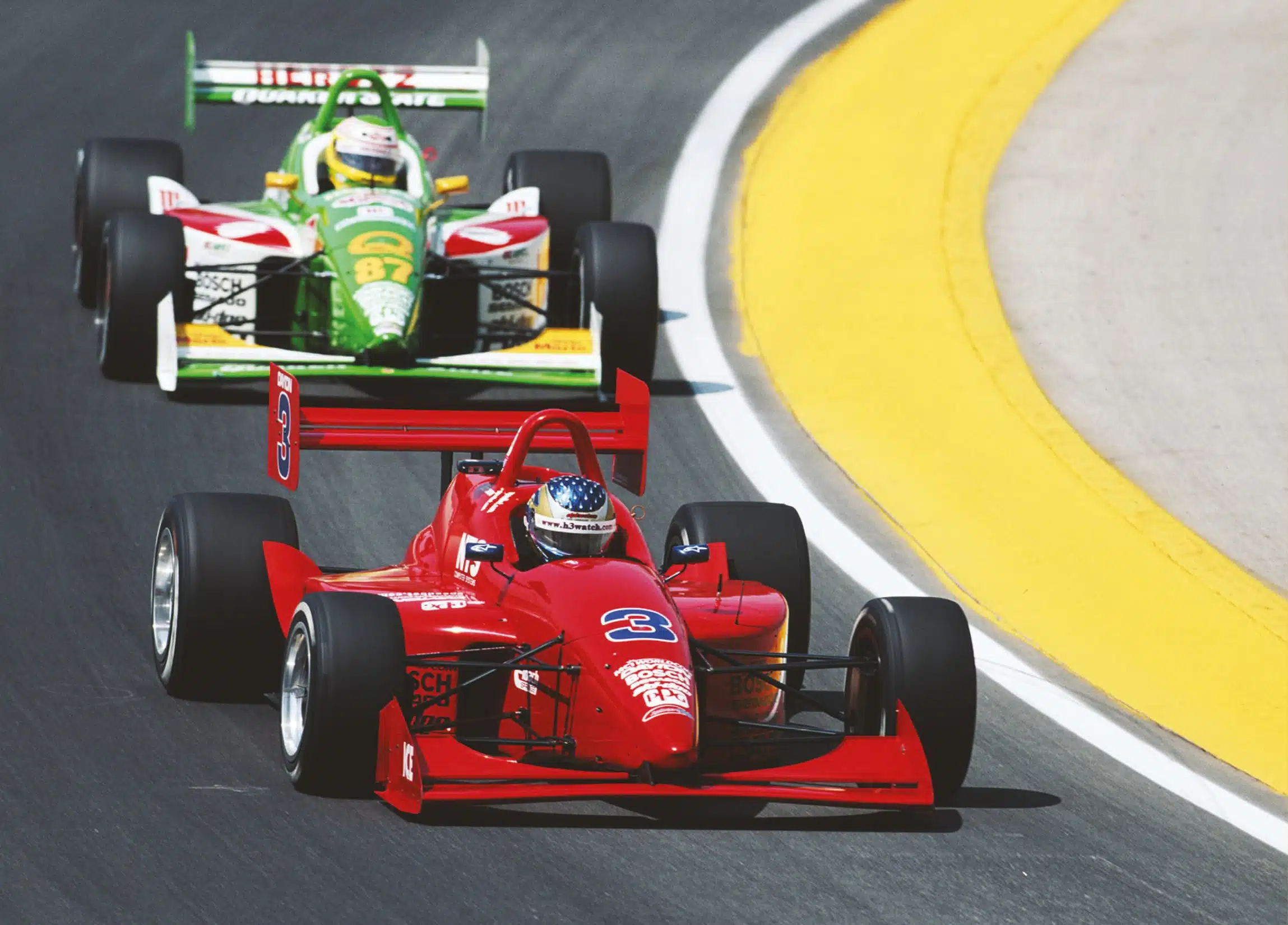 Red No.3 Bosch Goodyear IndyCar with American flag helmet leads green No.87 Quaker State car on curved racetrack